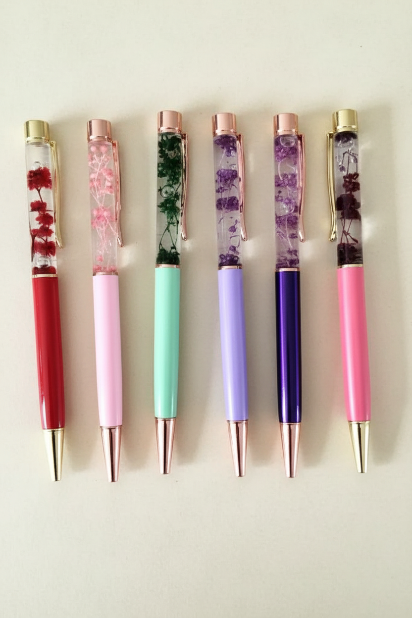 6 oil-filled dried flower ballpoint pens laid flat on a dark cream background